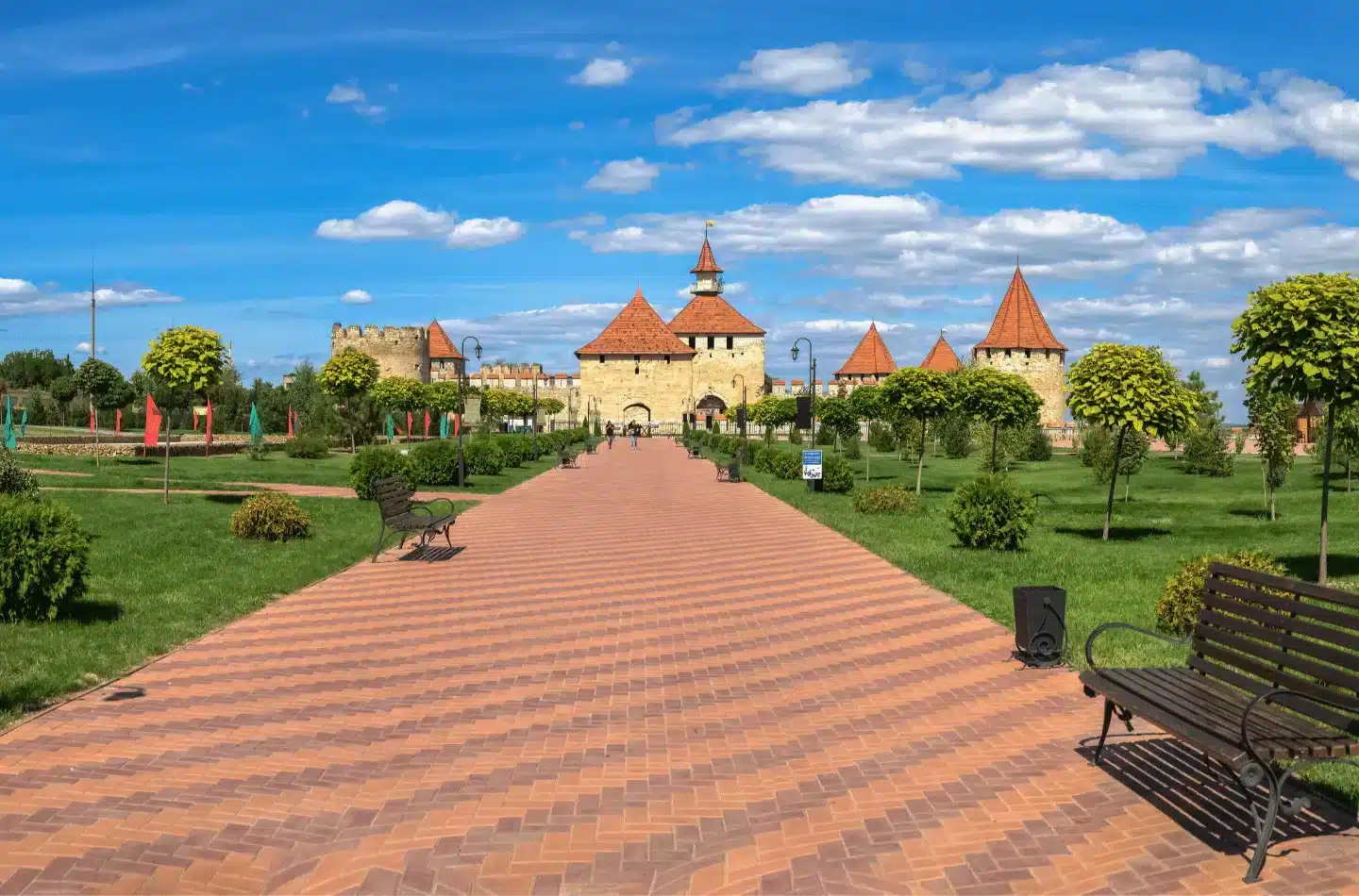 Wide walkway through a green park leading toward Bender Fortress with stone walls and red-roofed towers under a blue sky.